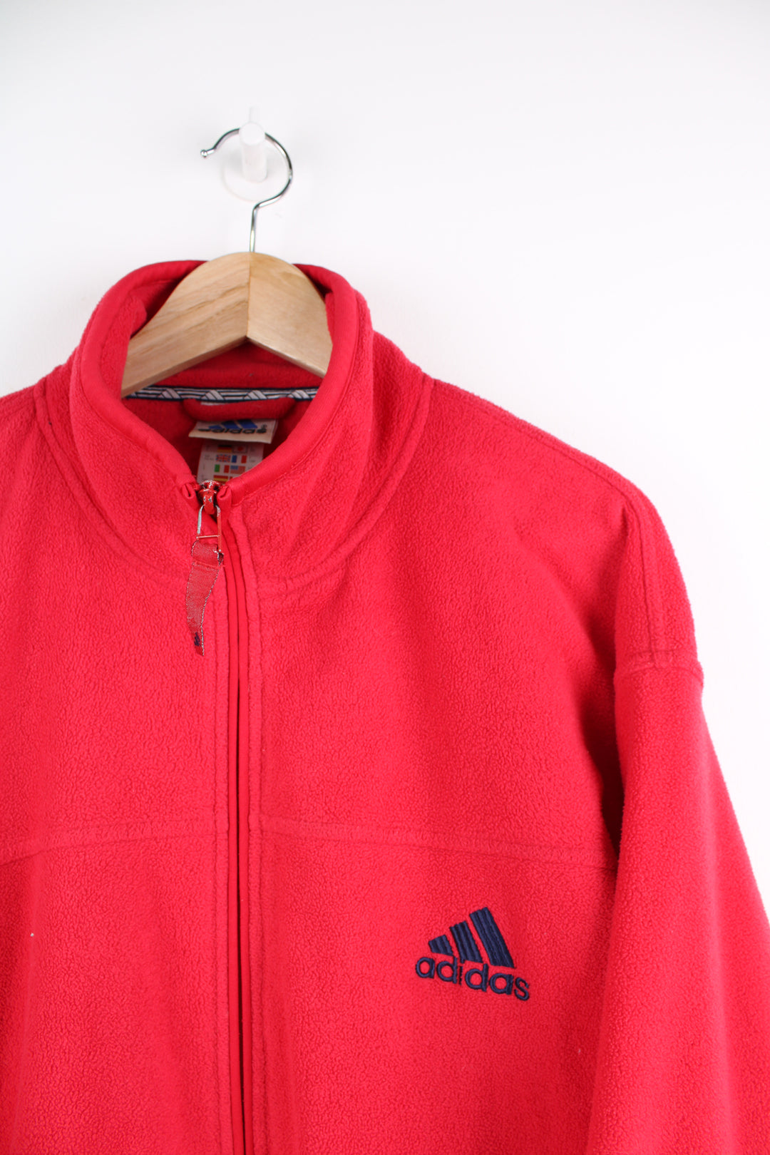 Adidas Fleece in a red colourway, navy blue iconic three stripes going down the sides, zip up, side pockets, and has the logo embroidered on the front.