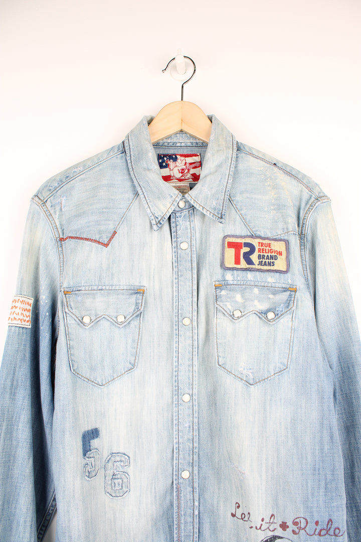 True Religion light wash heavily distressed denim shirt features embroidered badge and printed traditonal tattoo style graphics all over 