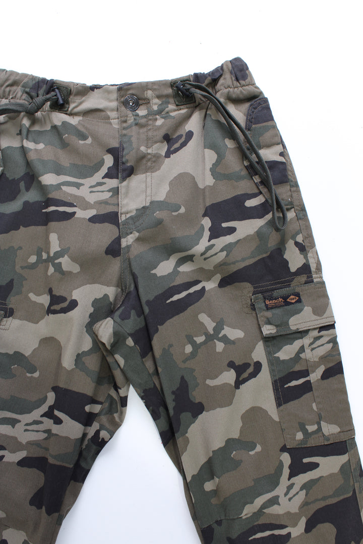 Bench Camo Cargo Trousers in a green, black and brown colourway, multiple utility style pockets, adjustable waist and cuffs, and has the logo embroidered on the front pocket.