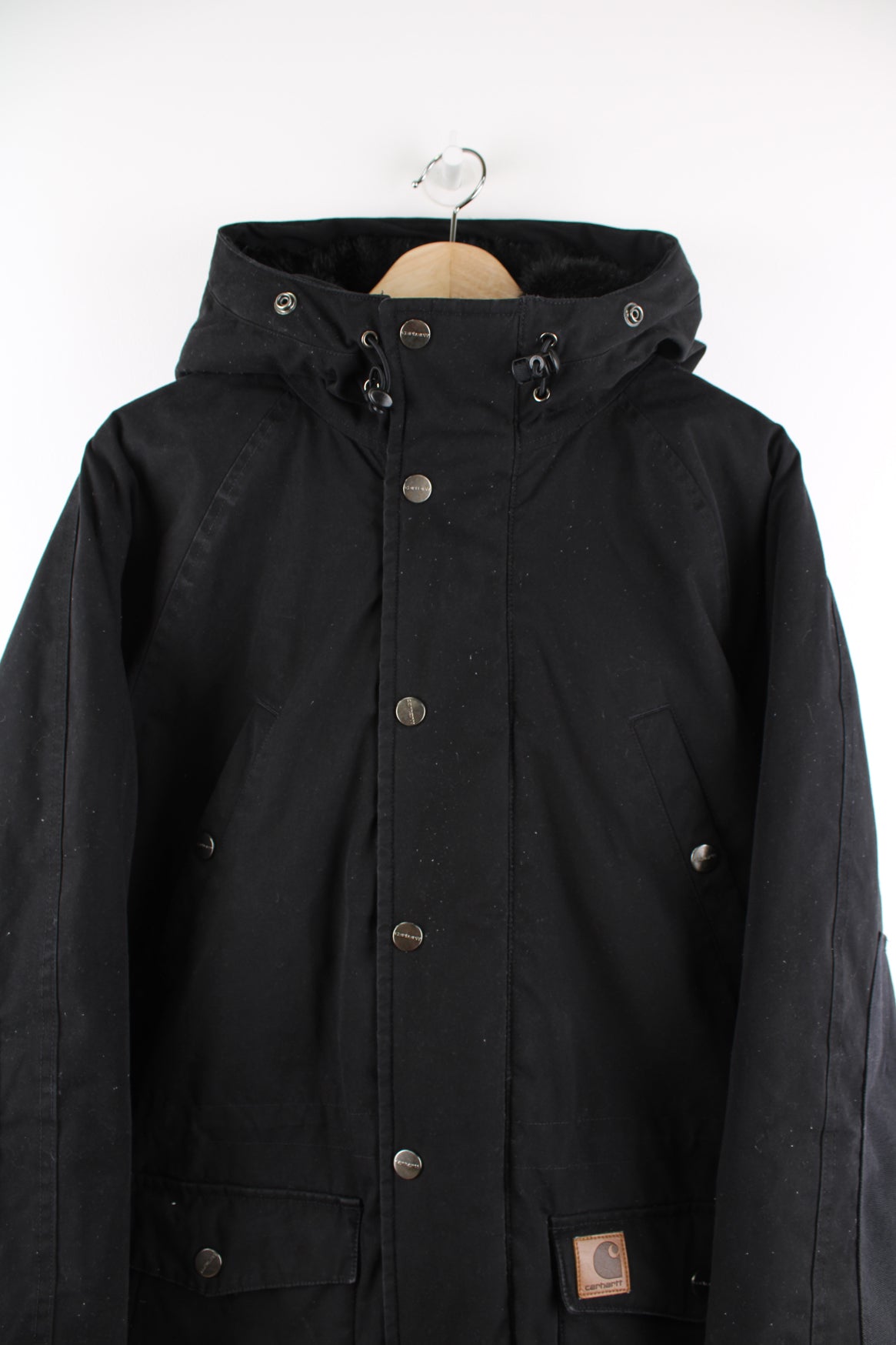 Carhartt Parka Jacket in a black colourway, zip up, multiple pockets, hooded with a faux fur lining and insulated with a quilted lining, and has the logo embroidered on the front.