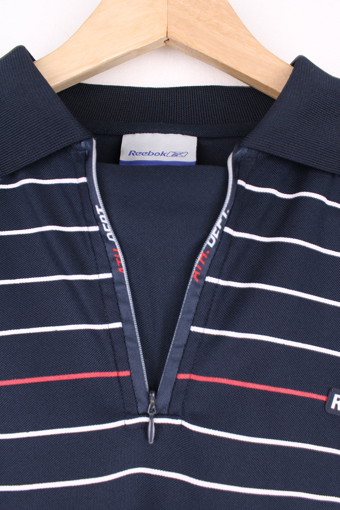 Reebok Athletic Polo Shirt in a navy blue colourway with white and red stripes going across the chest, zip up collar, short sleeved and has the logo spell out embroidered on the front.