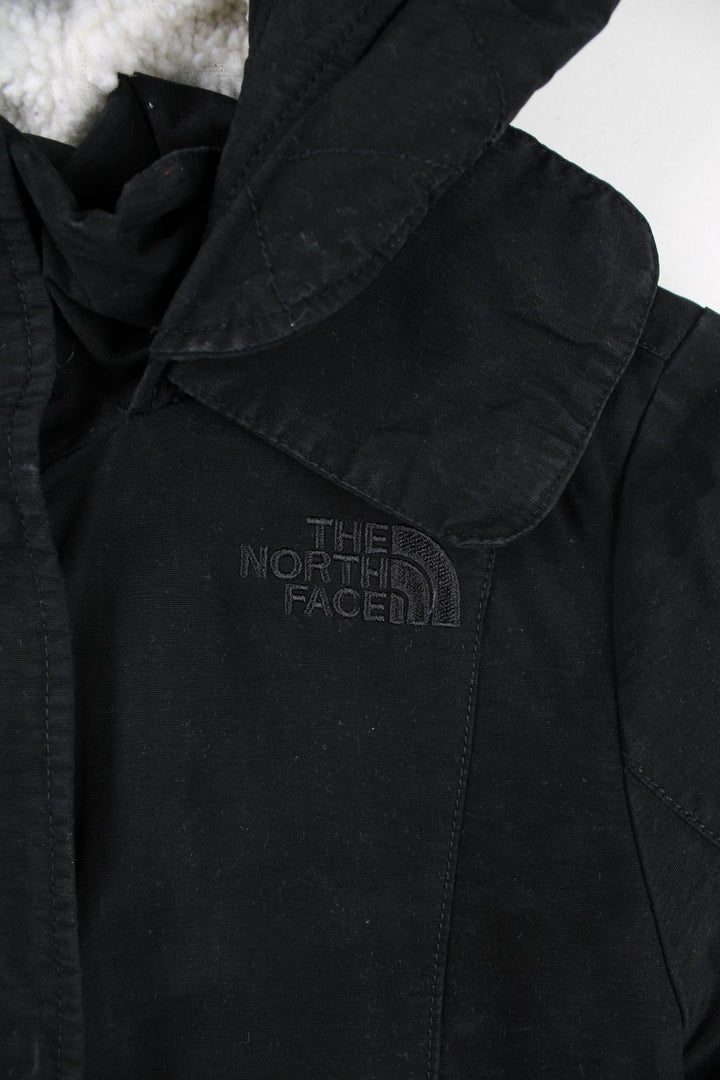 The North Face Parka Coat in a black colourway. Zips and buttons up, has side pockets, insulated with a sherpa lining, hooded, and has the logo embroidered on the front and back.