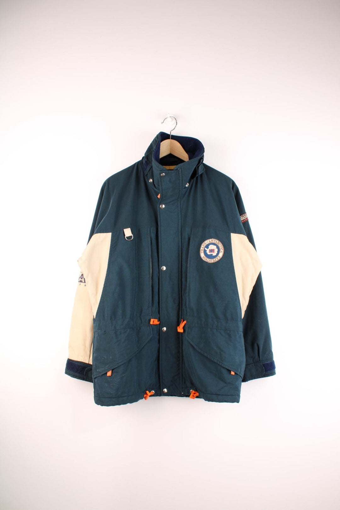 Napapijri Geographic Trans-Antarctica Expedition coat in navy blue and cream with a fleece-lined collar, zip and snap closure, multiple pockets, orange toggles, and patches including a logo on the sleeve and a Norway flag on the back.