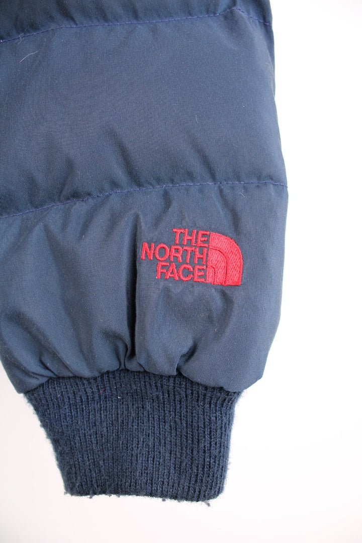 The North Face puffer coat in navy blue with red side-vents and lining, zip and snap closure, and an embroidered logo in red on the lower left sleeve.