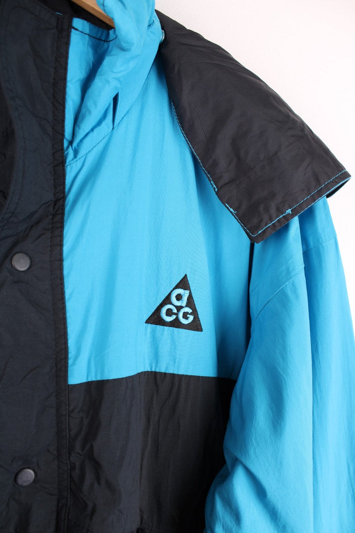 Nike ACG windbreaker jacket in black and blue with zip and snap closure, two zipped pockets, toggle adjusters, a detachable hood, and an embroidered triangle logo on the chest.