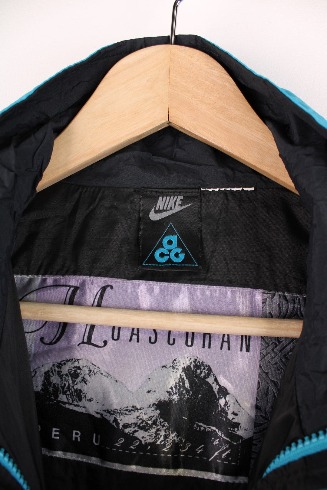 Nike ACG windbreaker jacket in black and blue with zip and snap closure, two zipped pockets, toggle adjusters, a detachable hood, and an embroidered triangle logo on the chest.