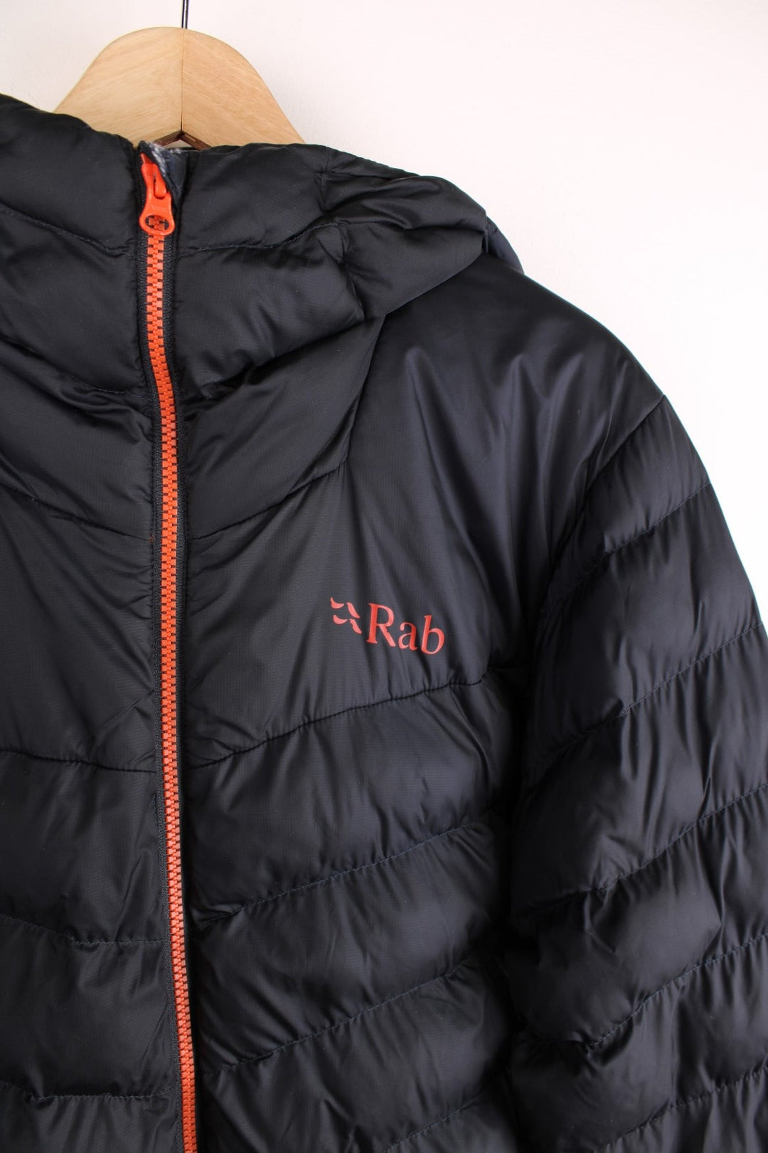 Rab puffer coat in black with orange accents and lining, zip closure, two zipped pockets, toggle adjusters, and a printed logo on the chest.