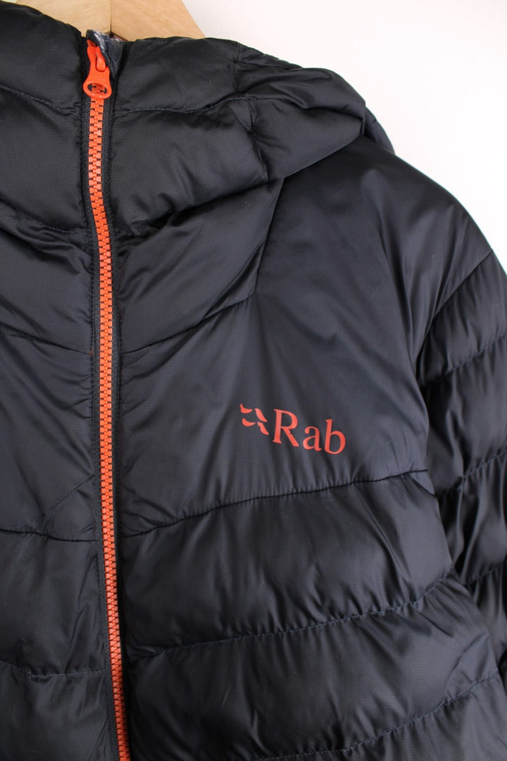 Rab puffer coat in black with orange accents and lining, zip closure, two zipped pockets, toggle adjusters, and a printed logo on the chest.