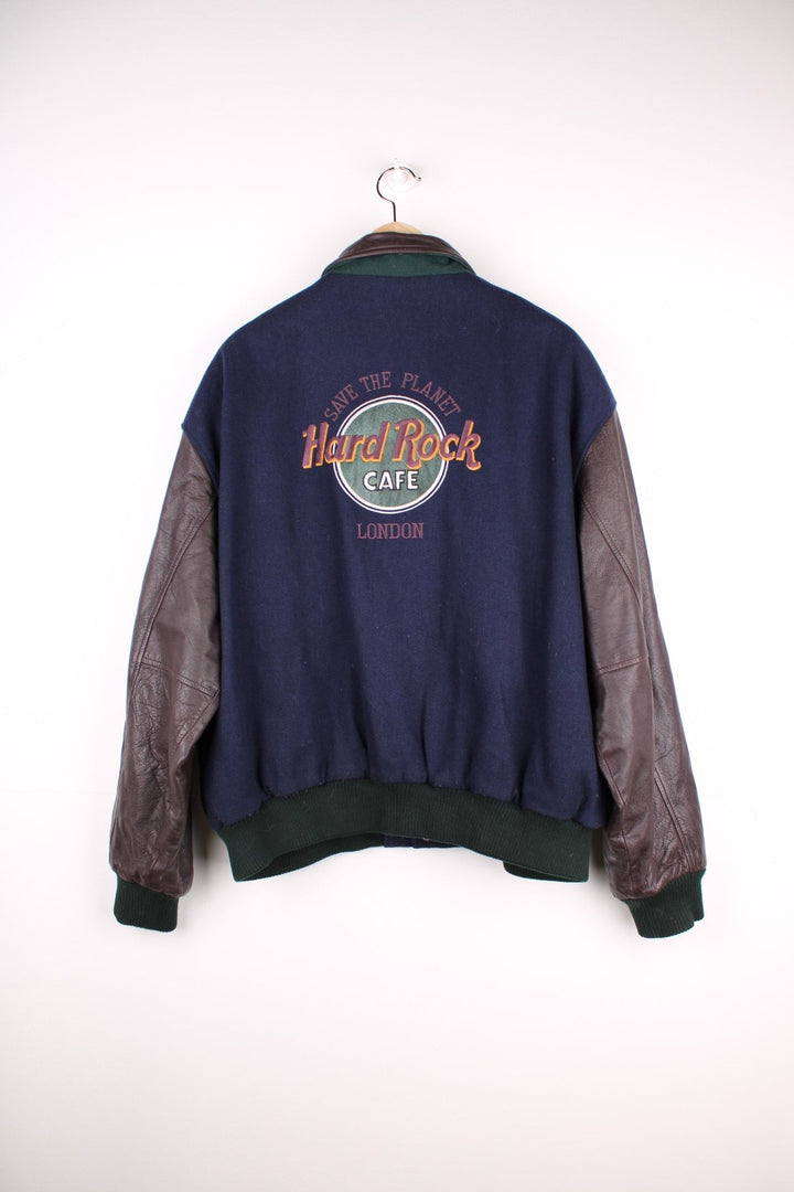 Vintage Hard Rock Cafe Jacket in a blue burgundy and green colourway. The jacket has leather sleeves, a green elasticated cuff and hem and Hard Rock logo press studs to close. On the back panel there is the - Hard Rock Cafe London - patch embroidery. The jacket also has one interior pocket and is lined with a quilted polyester filling.  