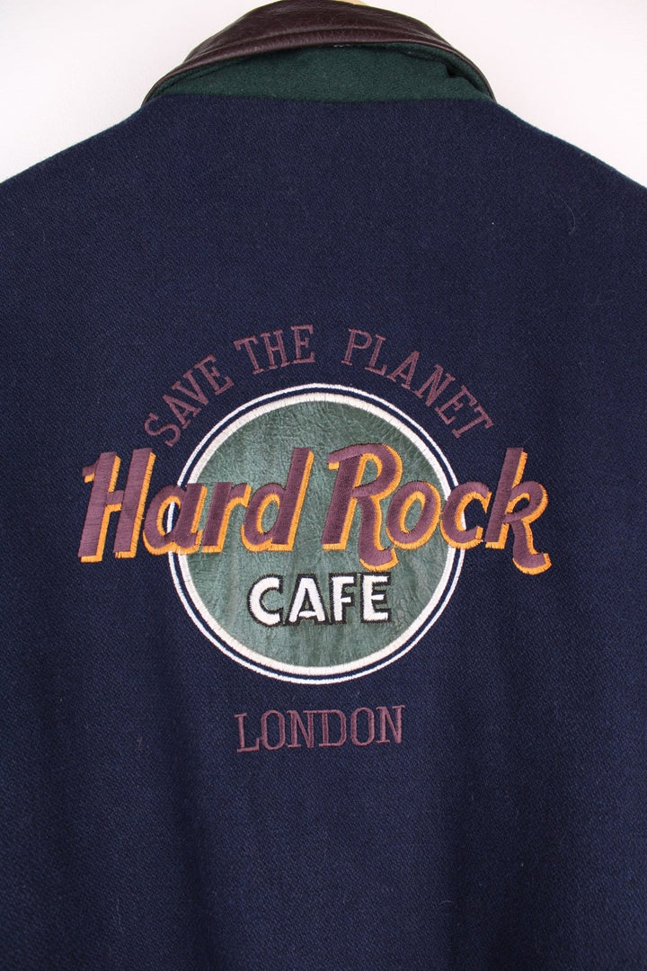 Vintage Hard Rock Cafe Jacket in a blue burgundy and green colourway. The jacket has leather sleeves, a green elasticated cuff and hem and Hard Rock logo press studs to close. On the back panel there is the - Hard Rock Cafe London - patch embroidery. The jacket also has one interior pocket and is lined with a quilted polyester filling.  