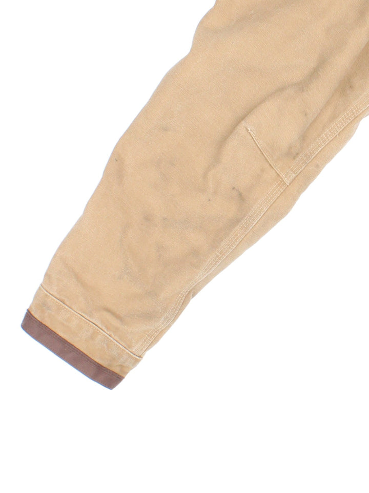 Carhartt J285 FRB Frontier Chore jacket in a tan colourway. The jacket has multiple pockets on the exterior with zip closures, and two interior pockets. There is a contrasting collar, and silver hardware. 