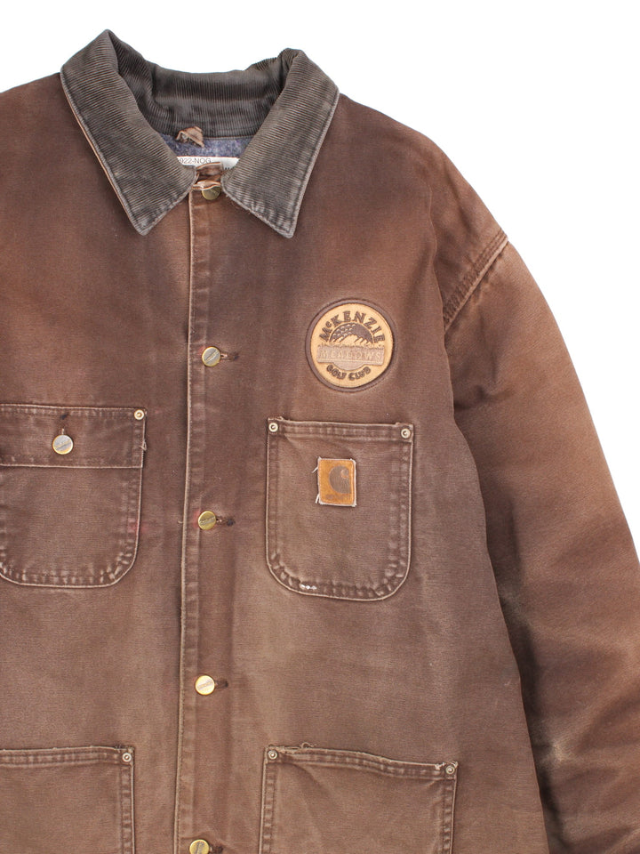 Vintage Carhartt Chore Coat in a brown colourway. The jacket has multiple exterior pockets, and one interior. It is blanket lined and has a corduroy collar. There is a leather logo patch stitched onto the front, and gold hardware.