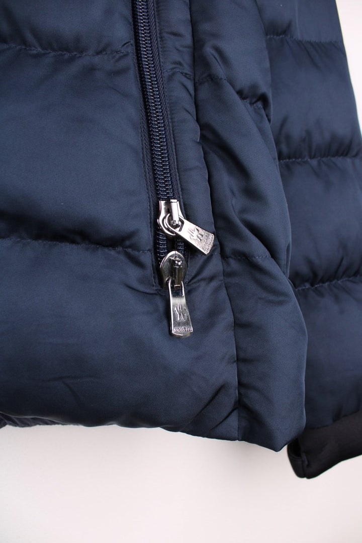 Moncler Cardere down puffer coat in a navy blue colourway. The coat has a puffer lining and the Moncler logo embroidered around the collar. There are extra thermal lined cuffs and a double zip for extra warmth.  