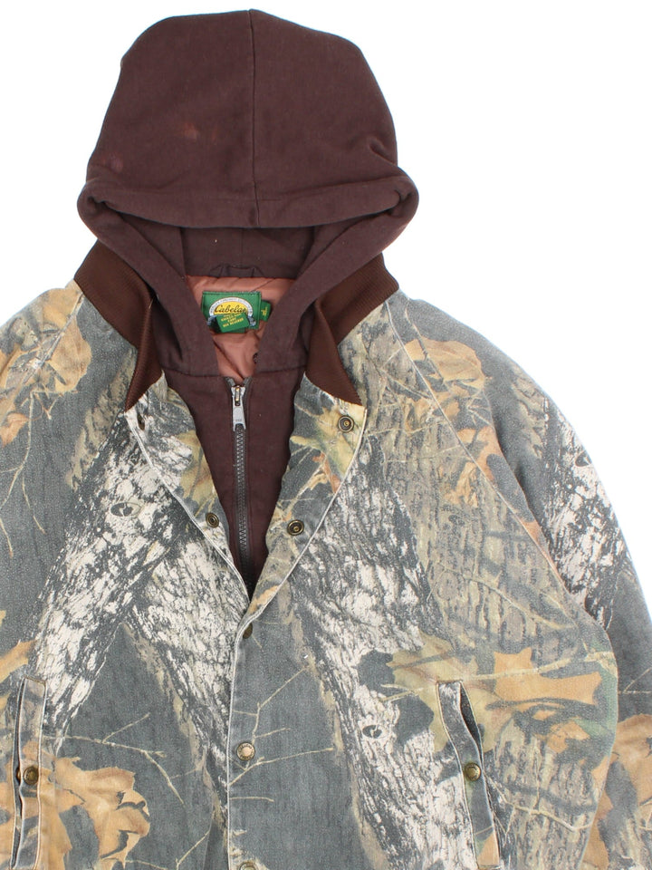 Vintage Cabela's Real Tree Camo Bomber Hunting Jacket in a greenand brown colourway. Zips and buttons up, has side pockets, insulated with a quilted lining, and hooded.