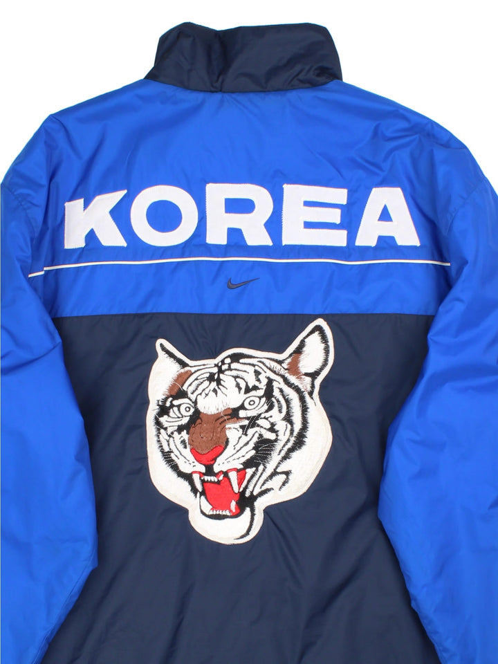 Vintage Nike Korea Coach Jacket in a blue colourway. Zips up and has side pockets, insulated with a quilted lining, and the logo embroidered on the front and on the back Korea spell out with embroidered tiger graphic.