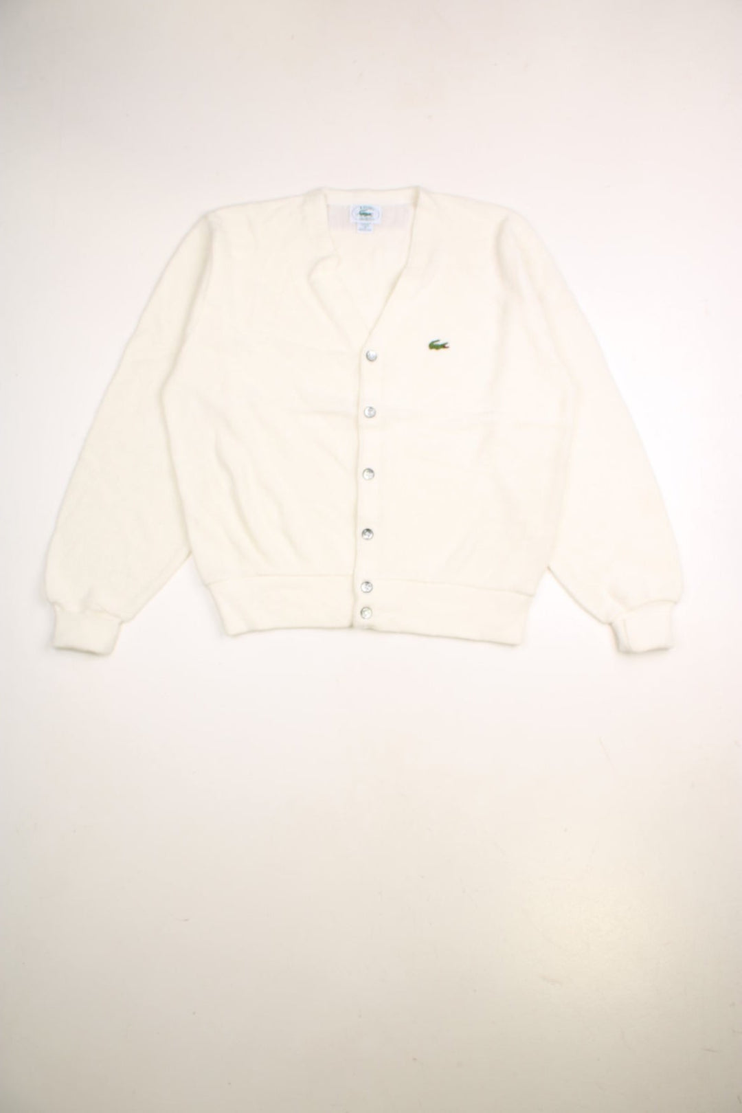 Lacoste cardigan in a off white /creamy colourway with the Lacoste logo stitched on the front. The cardigan has silvery grey buttons and cuffed sleeves. Made in USA. 