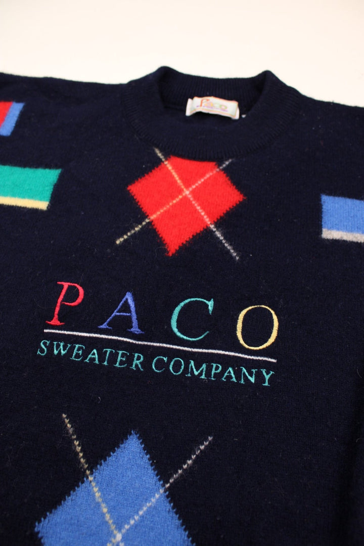 Paco patterned jumper in blue with geometric designs in bright primary colours, and an embroidered spellout reading: 'PACO Sweater Company'.