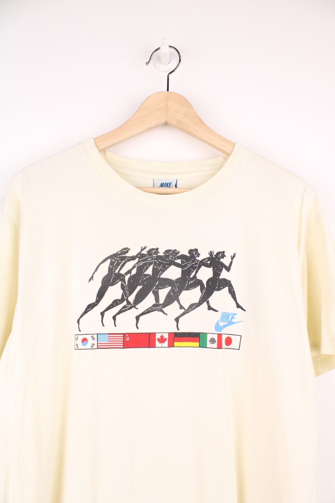 Vintage Nike 1988 Olympic Runners T-Shirt in a pale yellow colourway, has the runners and country flags graphic design printed on the front, as well as the logo and &