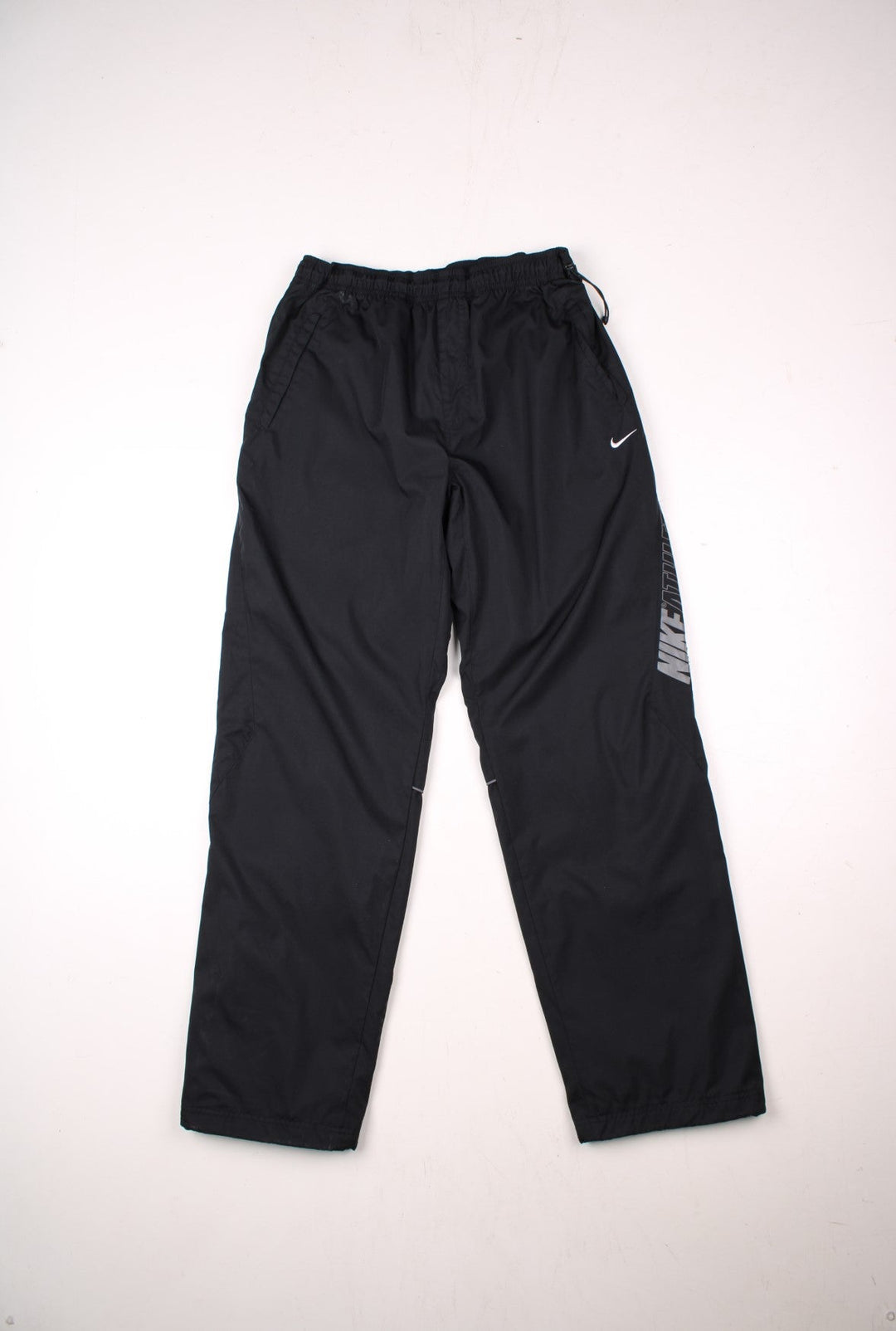 Nike Athletic 72 tracksuit bottoms in black with an elasticated waist, a toggle adjuster, two pockets, a printed spellout up the left leg, and an embroidered logo on the front.