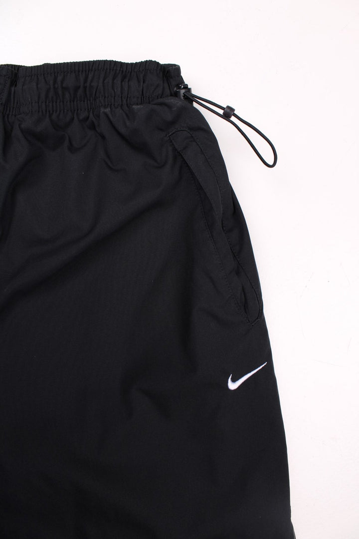 Nike Athletic 72 tracksuit bottoms in black with an elasticated waist, a toggle adjuster, two pockets, a printed spellout up the left leg, and an embroidered logo on the front.