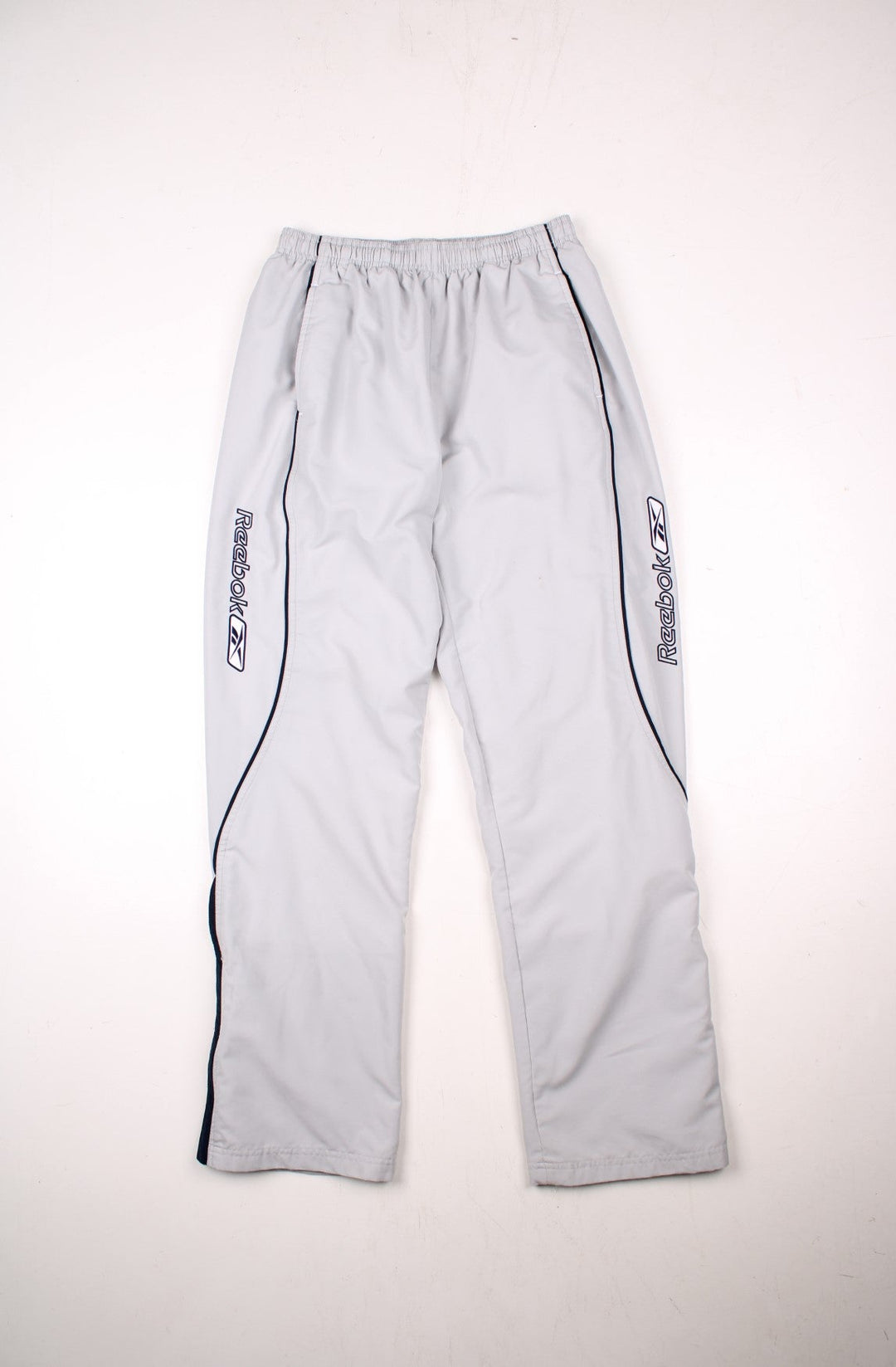 Reebok tracksuit bottoms in tan with navy blue piping, two pockets, an elasticated waist, and an embroidered logo along each leg.