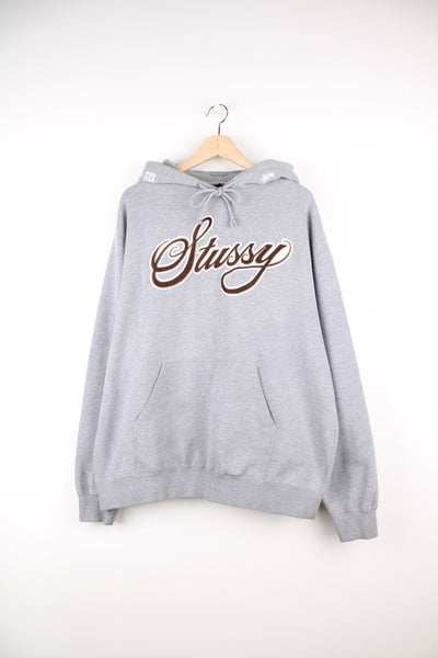 Stussy Logo Hoodie in a grey colourway with the spell out logo embroidered across the front in a brown and white colourway, also has 'XXV' embroidered onto the hood alongside the logo, and has a pouch pocket.