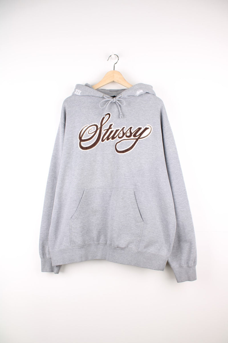 Stussy Logo Hoodie in a grey colourway with the spell out logo embroidered across the front in a brown and white colourway, also has 'XXV' embroidered onto the hood alongside the logo, and has a pouch pocket.