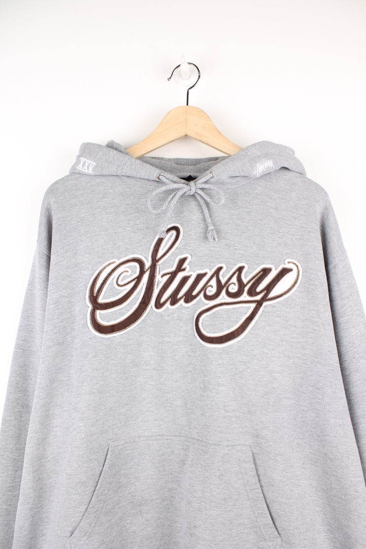 Stussy Logo Hoodie in a grey colourway with the spell out logo embroidered across the front in a brown and white colourway, also has 'XXV' embroidered onto the hood alongside the logo, and has a pouch pocket.