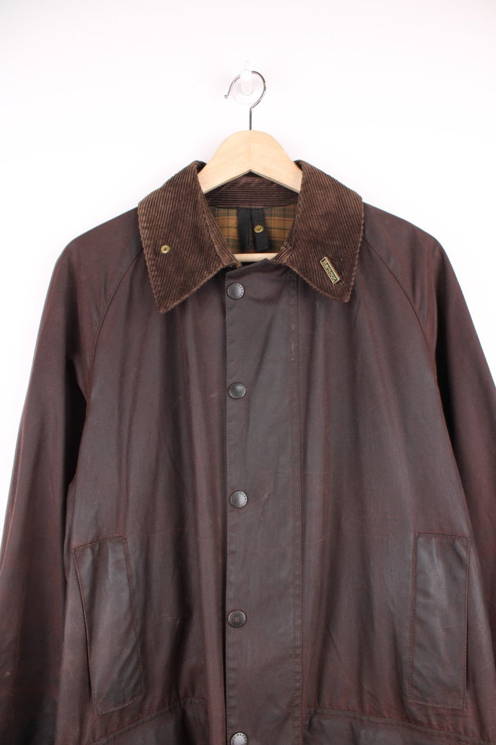 Vintage Barbour Beaufort Wax Jacket in a burgundy brown colourway with a corduroy collar that has a pin on it, zip up with multiple pockets, and has a tartan lining.