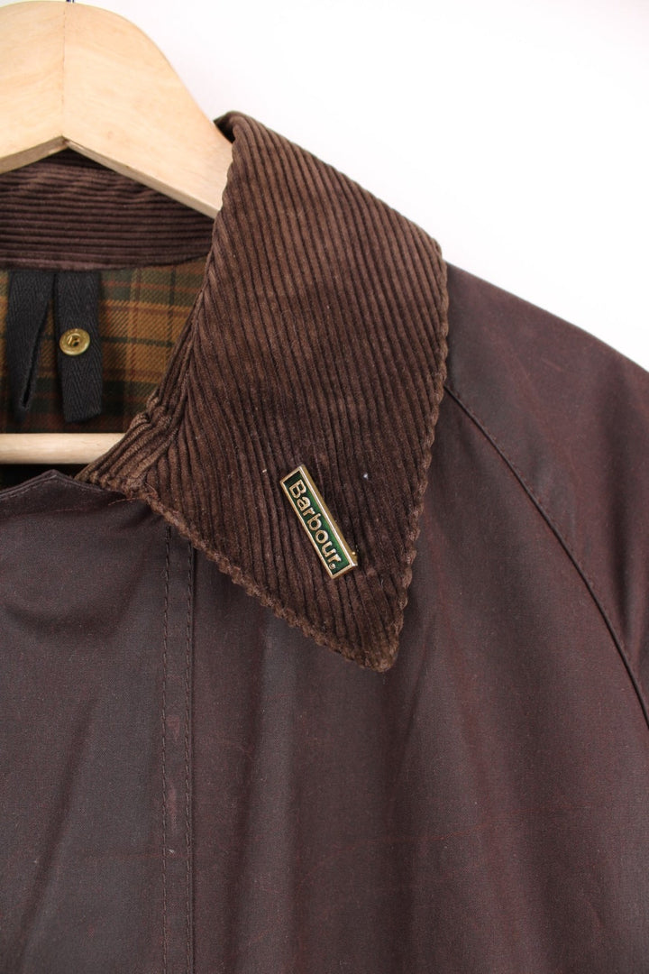 Vintage Barbour Beaufort Wax Jacket in a burgundy brown colourway with a corduroy collar that has a pin on it, zip up with multiple pockets, and has a tartan lining.