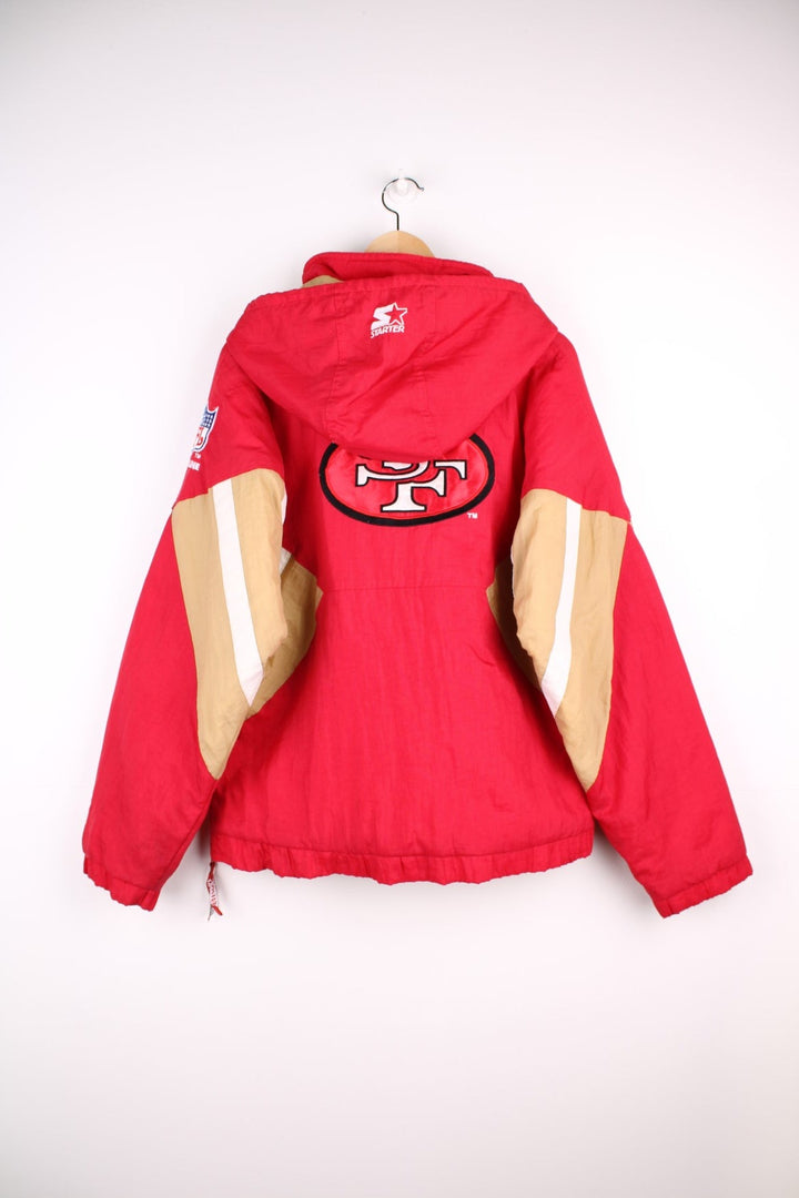 San Francisco 49ers, NFL pro sport jacket in a red colourway with beige and white side panels. Zip closure and hood, with team logos on the front, sleeve and back.