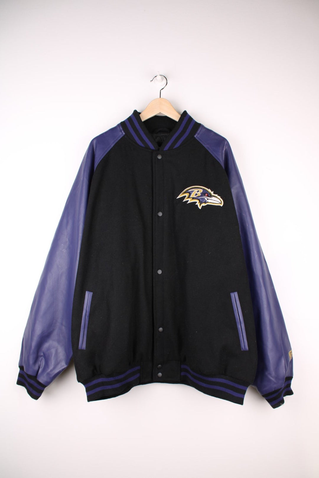 Baltimore Ravens NFL Varsity Jacket in a black and purple colourway, button up with side pockets, insulated with a quilted lining, and has the team logo and spell out embroidered on the front and back. 