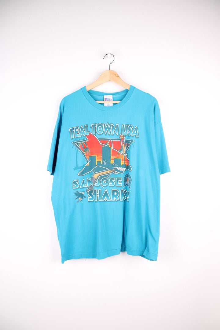 San Jose Sharks NHL t-shirt in a turquoise blue with spellout reading 'Teal Town USA, San Jose Sharks', front graphic of shark in orange, and small logo embroidered on the sleeve.