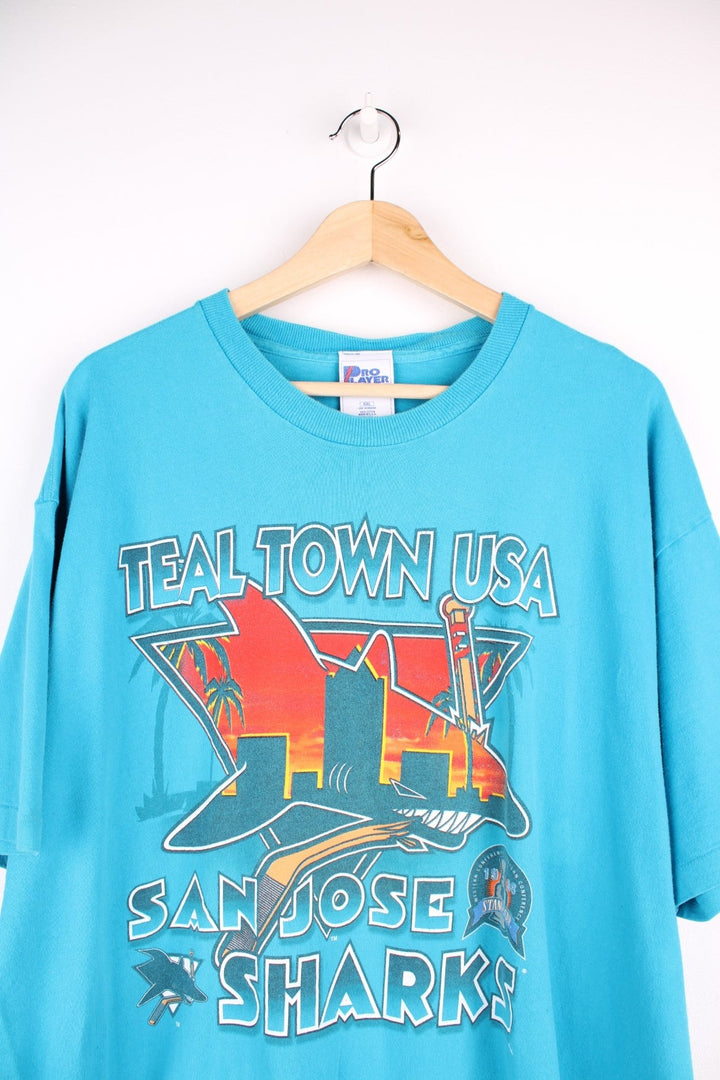 San Jose Sharks NHL t-shirt in a turquoise blue with spellout reading 'Teal Town USA, San Jose Sharks', front graphic of shark in orange, and small logo embroidered on the sleeve.