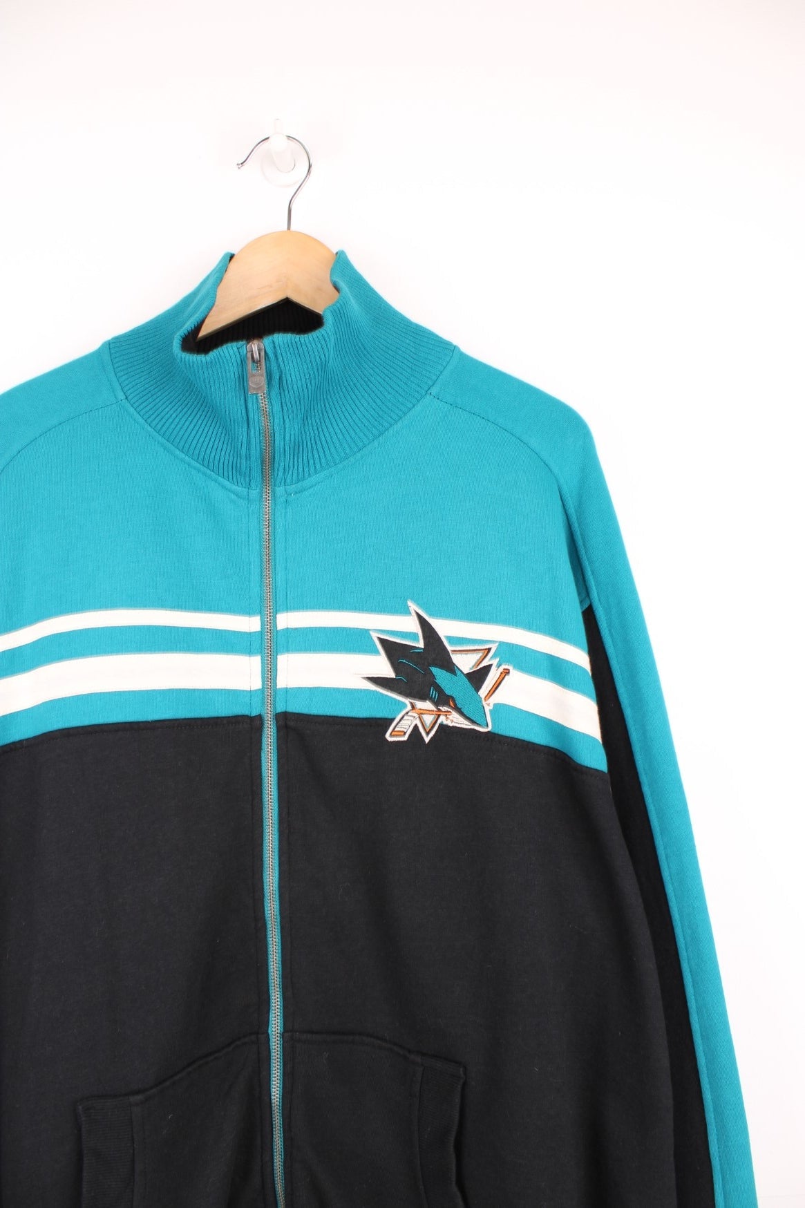 San Jose Sharks Zip Up Sweater in a black, blue and white colourway, full zip up with side pockets, stripes going across the front and the logo and spell out embroidered on the back.