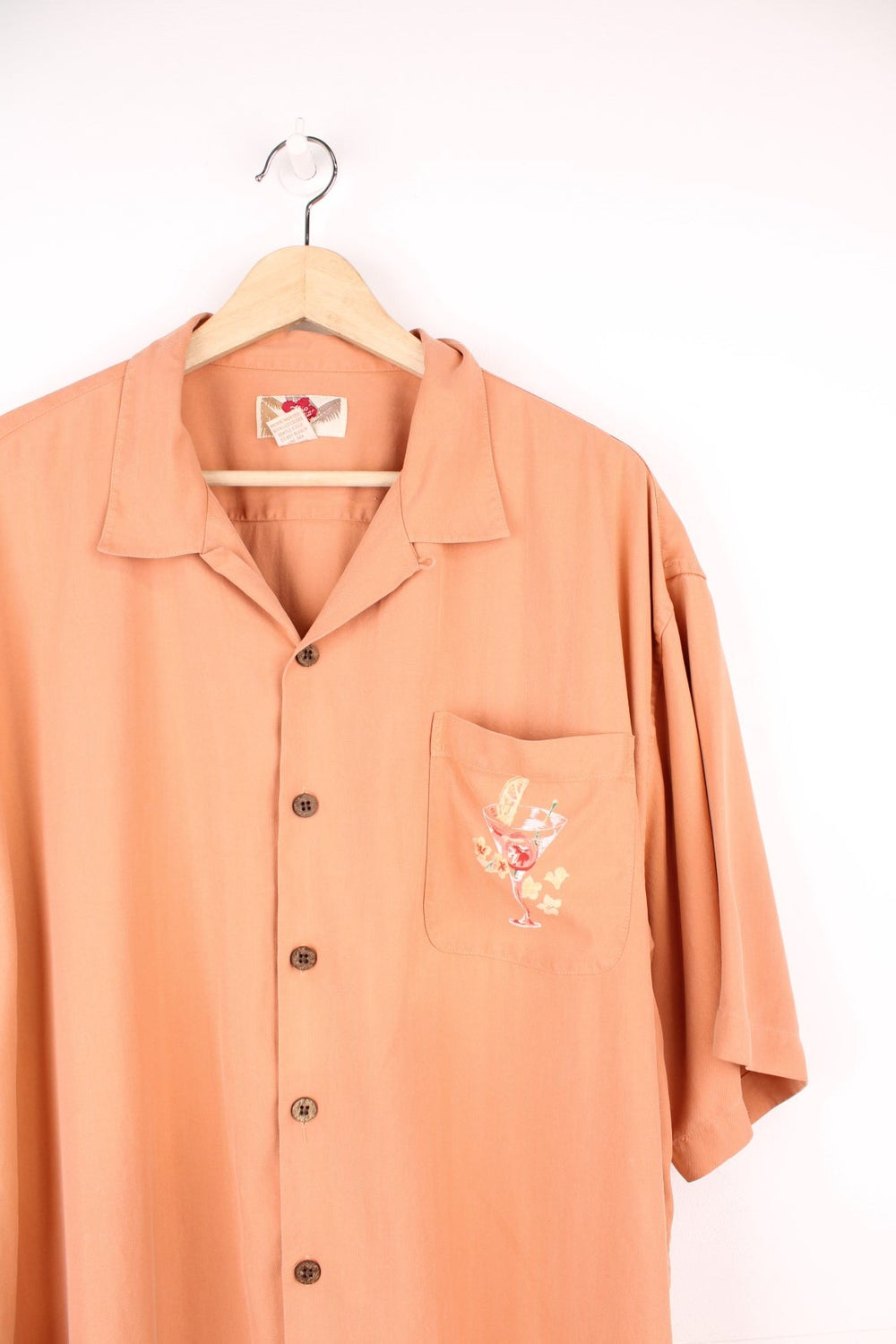 Peachy orange Hawaiian shirt by Hilo Hattie featuring tropical cocktail glass designs on the breast pocket and back of the shirt, and brown wooden buttons. 