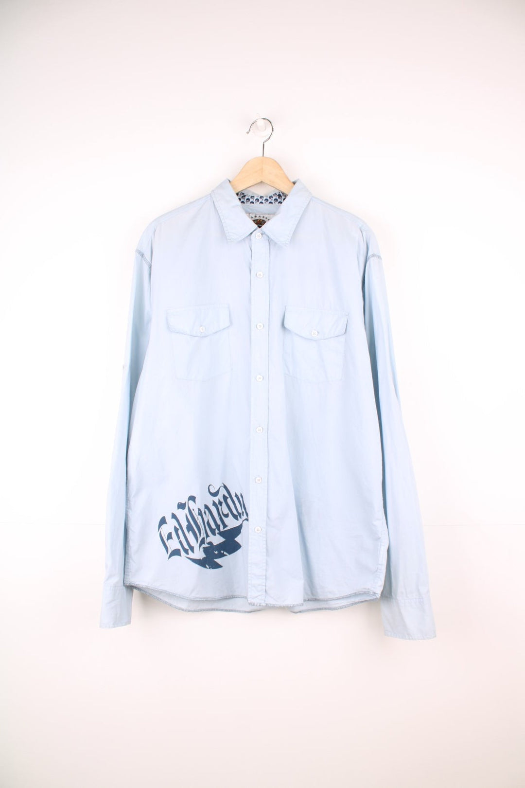 Pale blue long-sleeved button-down shirt by Ed Hardy, featuring double flap pockets, Ed Hardy spellout, tattoo-style eagle, tiger, roses and flames design on the back, skull motif on the collar and cuff lining, and an attached red brand tab on the left sleeve. 