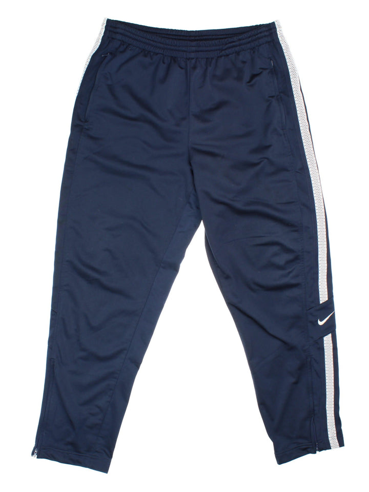 Nike tracksuit bottoms in navy blue with white tape detailing, an elasticated waist, two zipped pockets, and embroidered swoosh logos in white.