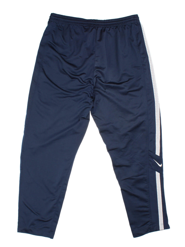 Nike tracksuit bottoms in navy blue with white tape detailing, an elasticated waist, two zipped pockets, and embroidered swoosh logos in white.