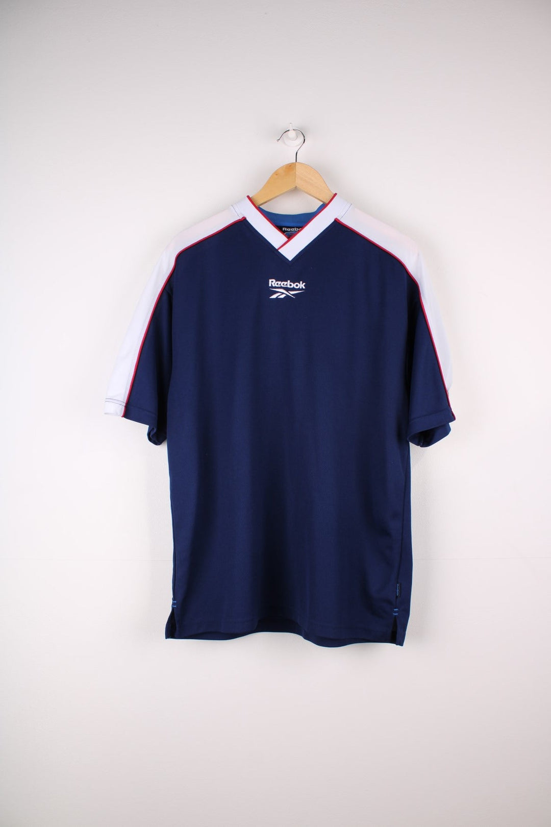 Vintage Reebok T-Shirt in a navy blue colourway. The shirt has a small Reebok logo embroidered on the front and red piping over the shoulders and collar. There is also a logo patch stitched on the arm. 