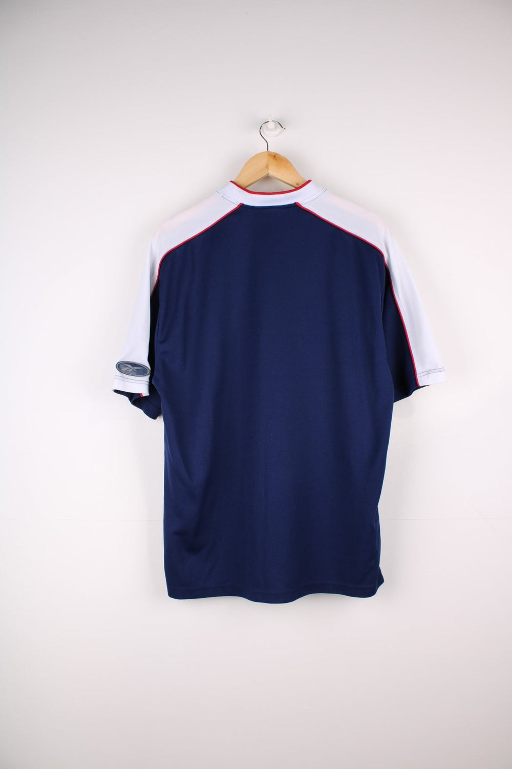 Vintage  T-Shirt in a navy  colourway. The shirt has a small Reebok logo embroidered on the front and red piping over the shoulders and collar. There is also a logo patch stitched on the arm. 