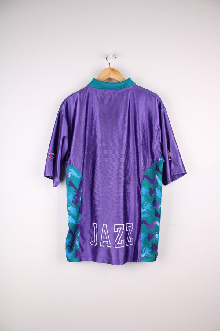 Vintage Champion Utah Jazz polo shirt in a purple colourway with a turquoise collar, and zip closure. The shirt has patterned side panels and the Utah Jazz logo embroidered on the front. There is also a Champion NBA authenticity patch sewn on the hem.