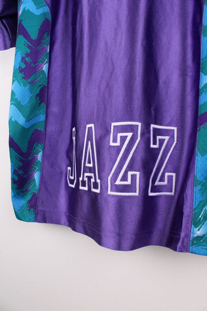 Vintage Champion Utah Jazz polo shirt in a purple colourway with a turquoise collar, and zip closure. The shirt has patterned side panels and the Utah Jazz logo embroidered on the front. There is also a Champion NBA authenticity patch sewn on the hem.