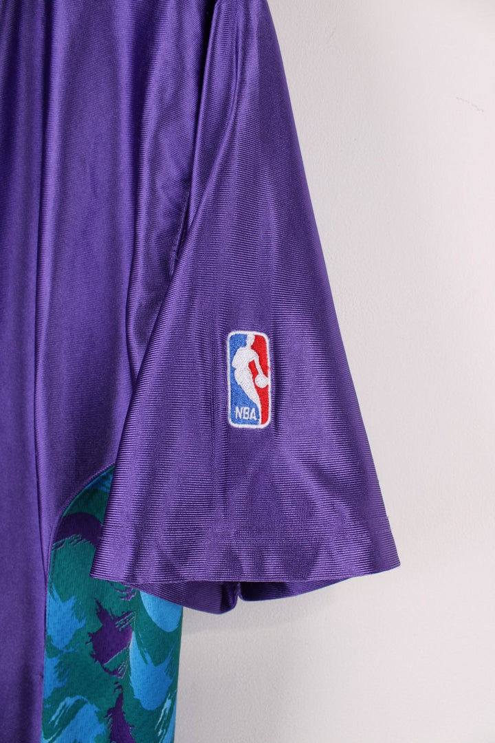 Vintage Champion Utah Jazz polo shirt in a purple colourway with a turquoise collar, and zip closure. The shirt has patterned side panels and the Utah Jazz logo embroidered on the front. There is also a Champion NBA authenticity patch sewn on the hem.