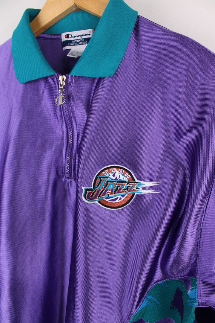 Vintage Champion Utah Jazz polo shirt in a purple colourway with a turquoise collar, and zip closure. The shirt has patterned side panels and the Utah Jazz logo embroidered on the front. There is also a Champion NBA authenticity patch sewn on the hem.