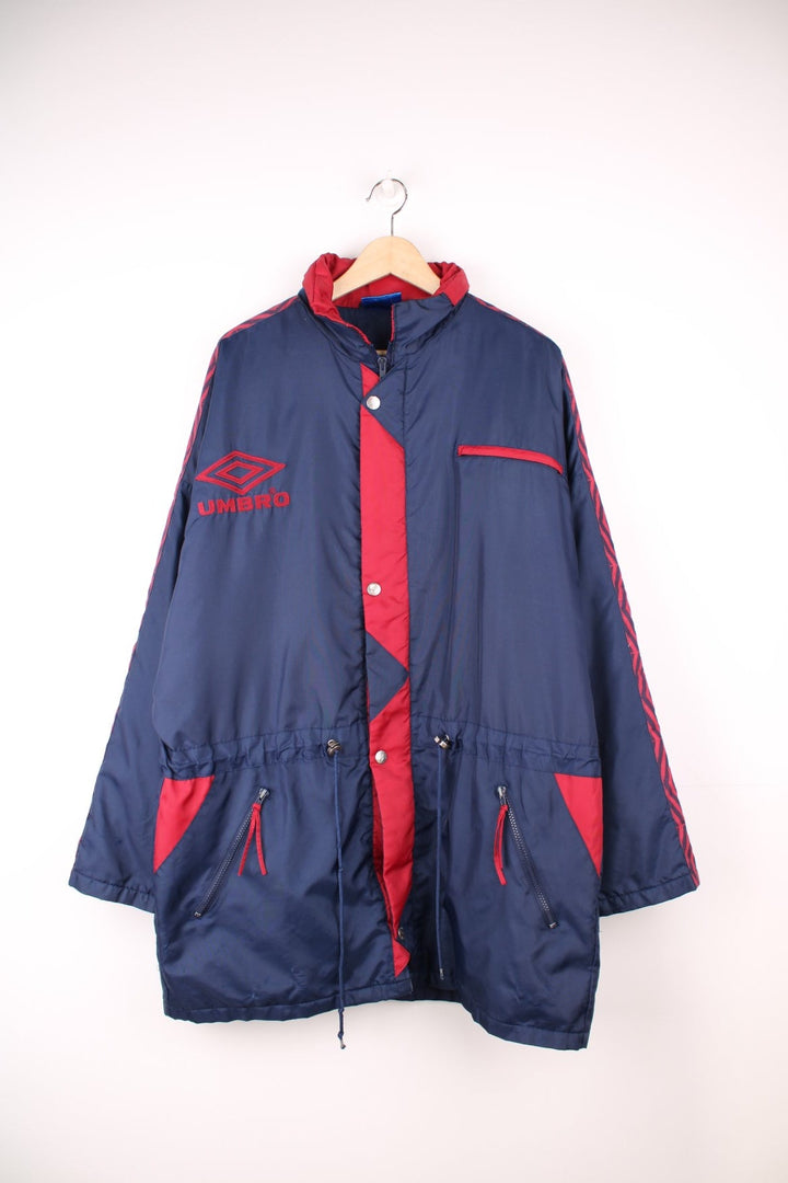 Umbro Training Coat in a navy blue and red colourway, zip up with multiple pockets, hidden hood, insulated, and has the logo embroidered on the front. 