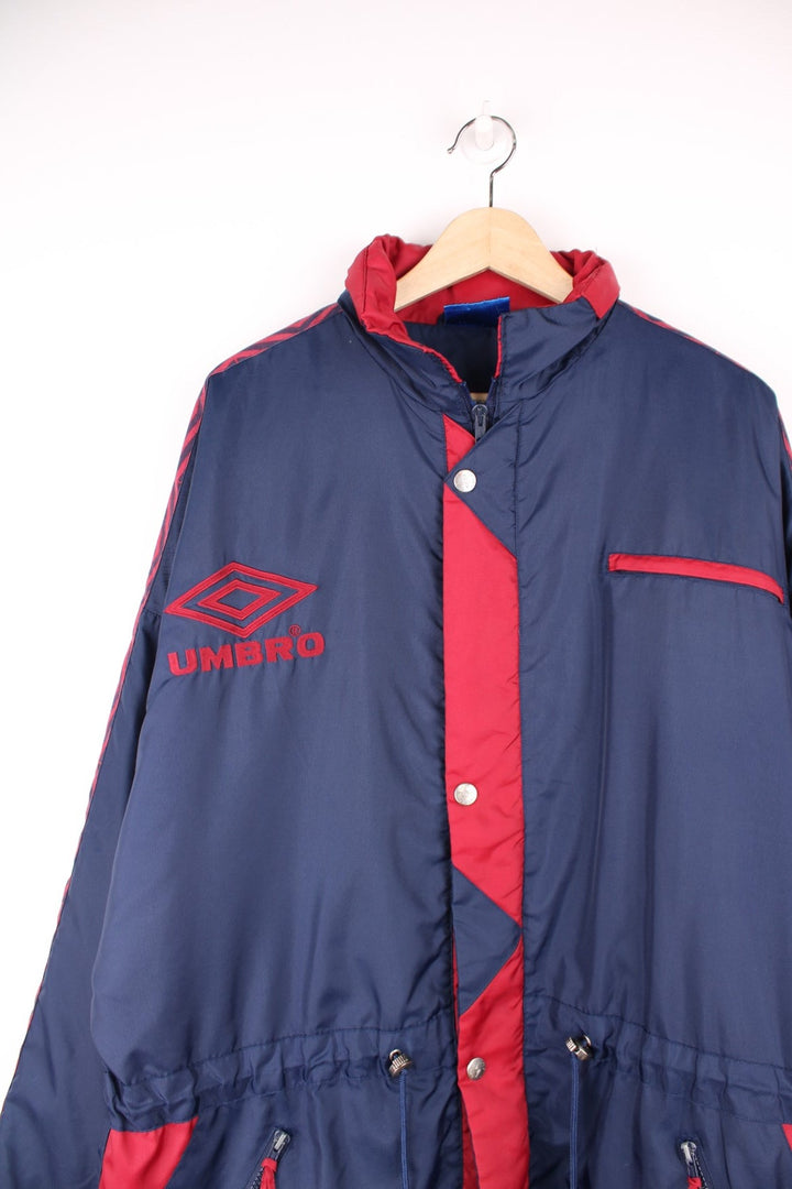 Umbro Training Coat in a navy blue and red colourway, zip up with multiple pockets, hidden hood, insulated, and has the logo embroidered on the front. 