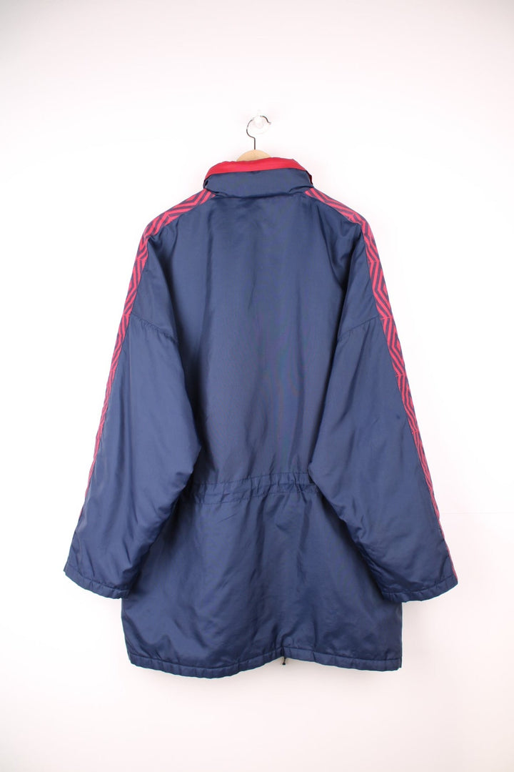 Umbro Training Coat in a navy blue and red colourway, zip up with multiple pockets, hidden hood, insulated, and has the logo embroidered on the front. 