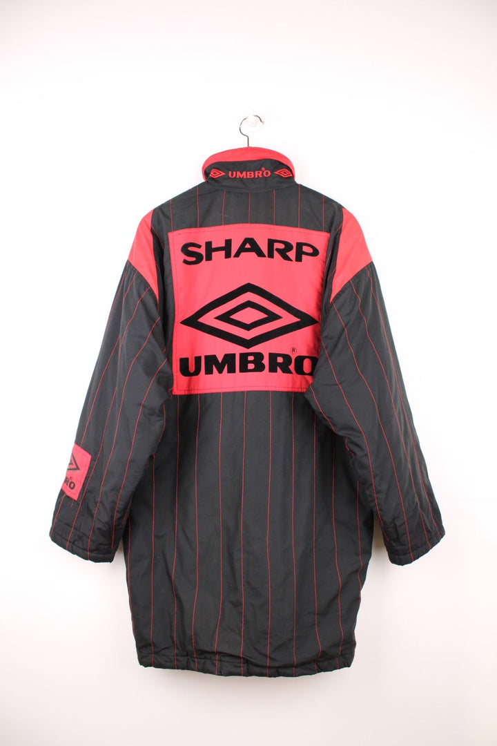 1993/95 Manchester United Umbro Training Coat in a black and red colourway with stripes throughout, zip up with side pockets, insulated with a quilted lining, and has the logos embroidered on the front and back.