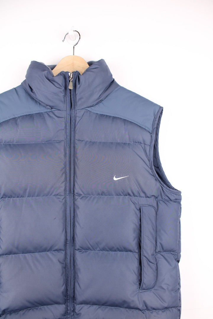 Nike Puffer Gilet in a blue colourway, zip up with side pockets, insulated lining, hidden hood, and has the swoosh logo embroidered on the front. 